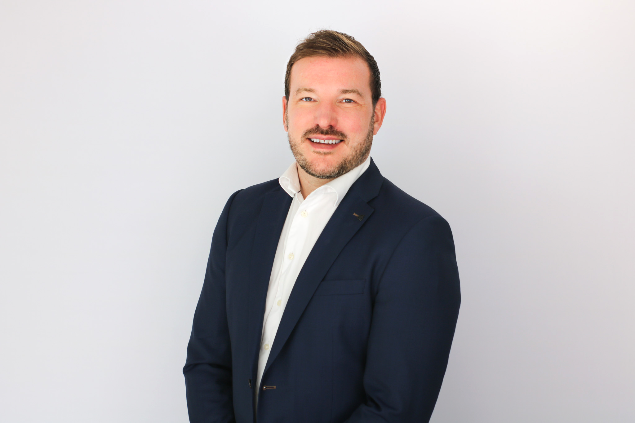 A smiling man with brown hair and facial hair wearing a dark navy blazer over a white shirt, photographed against a light gray background in a professional headshot style.