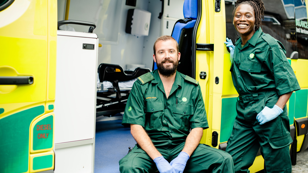 Two paramedics in green uniforms smile while posing with their yellow and green ambulance, one sitting in the driver's seat and the other standing beside the open door.