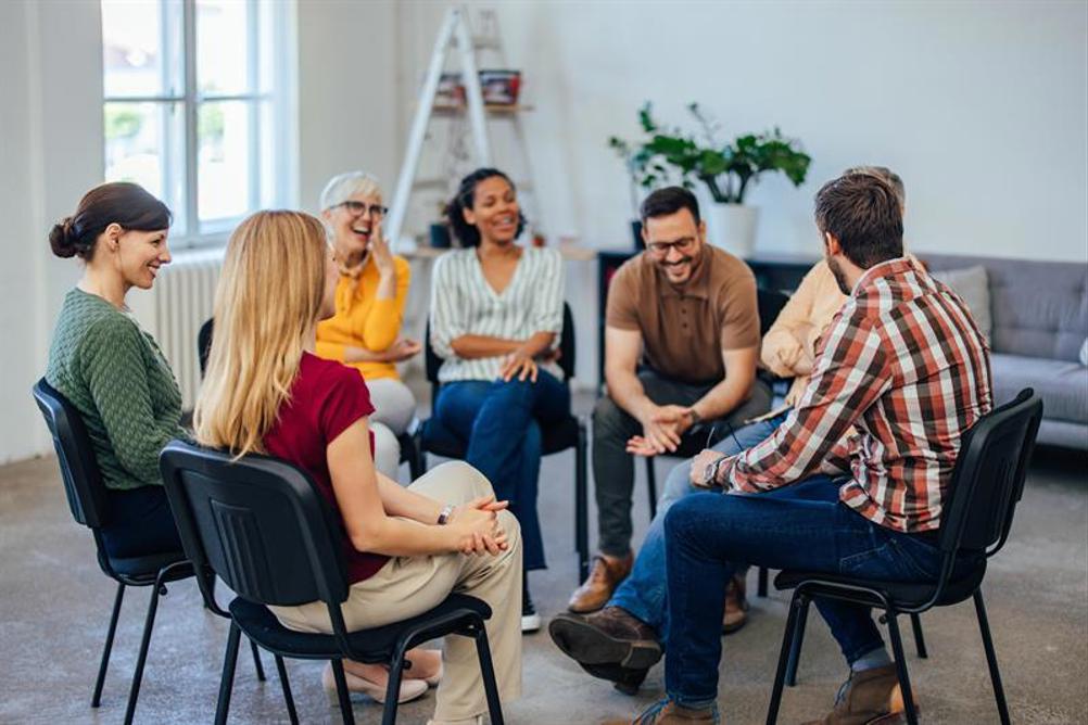 A diverse group of six people sit in a circle on black chairs in a bright, modern room, appearing to engage in a friendly group discussion or support meeting.