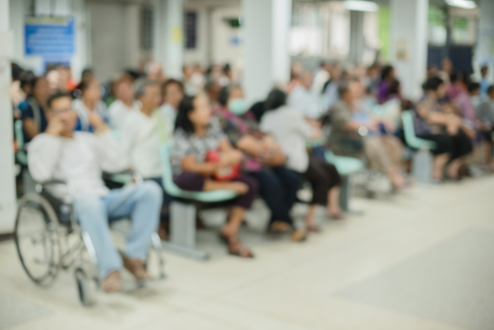 A crowded hospital or medical facility waiting area with people seated in chairs, including a person in a wheelchair in the foreground, all waiting to be seen by healthcare providers.