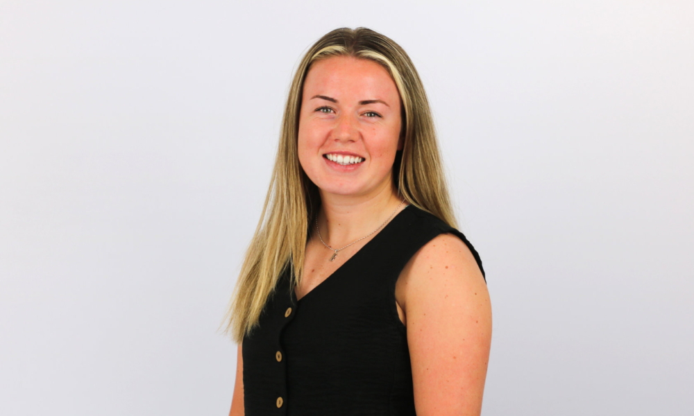 A smiling woman with long blonde hair wearing a black sleeveless top with decorative buttons poses against a light gray background in what appears to be a professional headshot.
