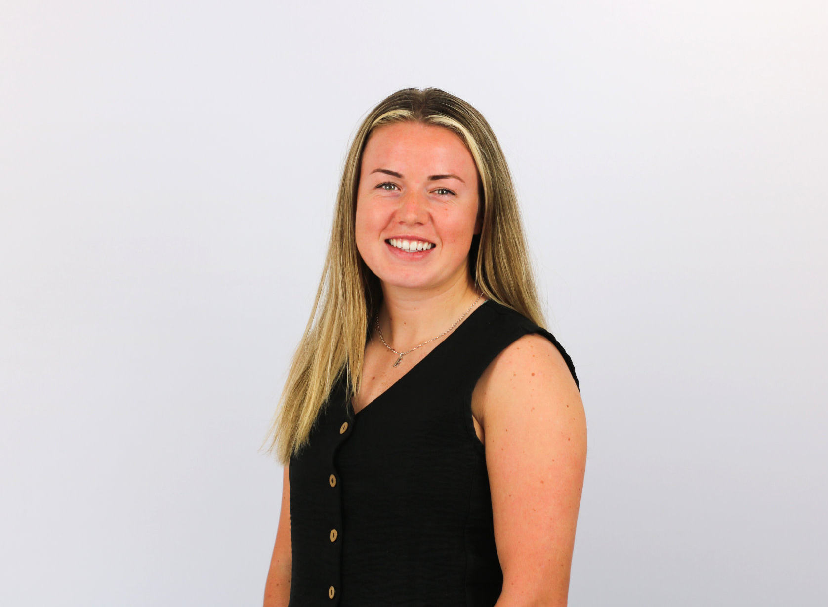 A smiling woman with long blonde hair wearing a black sleeveless top with decorative buttons poses against a light gray background in what appears to be a professional headshot.