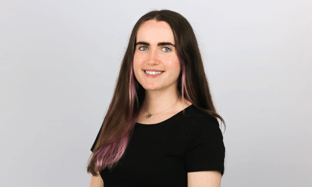 A young woman with long brown hair featuring purple highlights smiles at the camera while wearing a black short-sleeved top and small necklace against a light gray background.