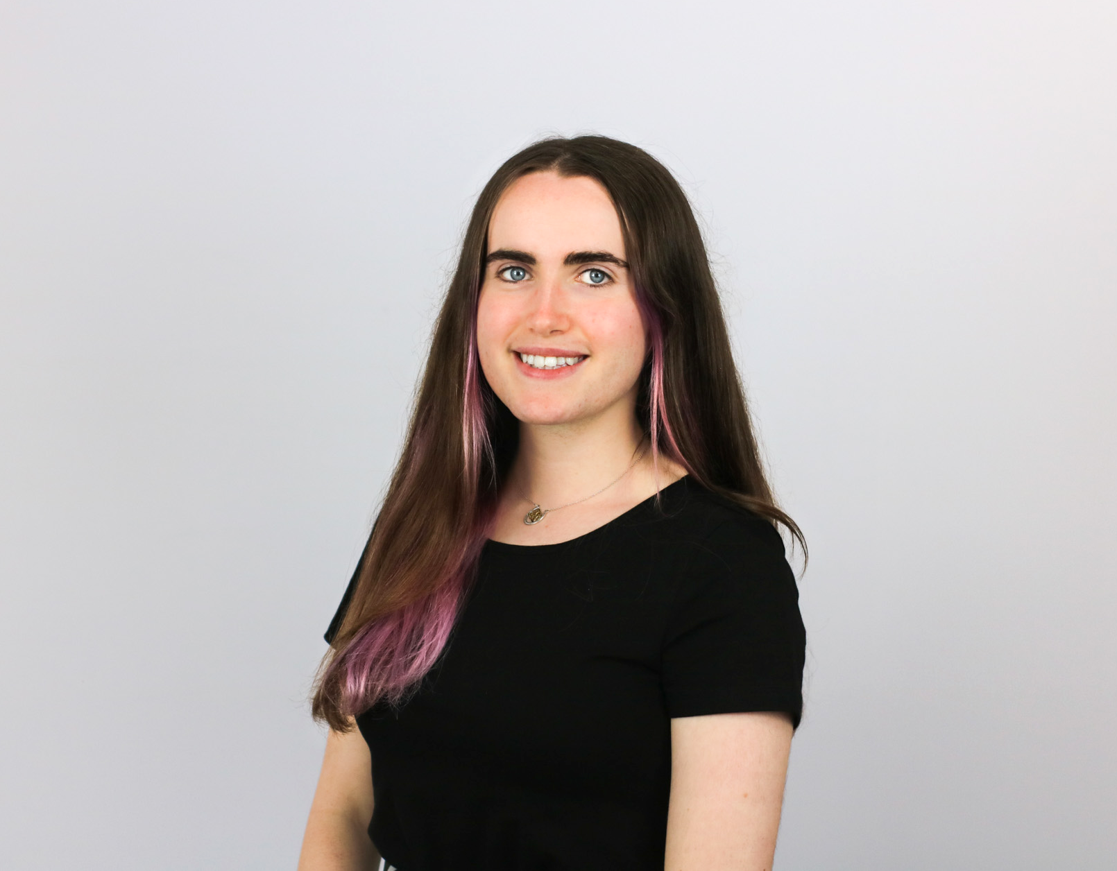 A young woman with long brown hair featuring purple highlights smiles at the camera while wearing a black short-sleeved top and small necklace against a light gray background.