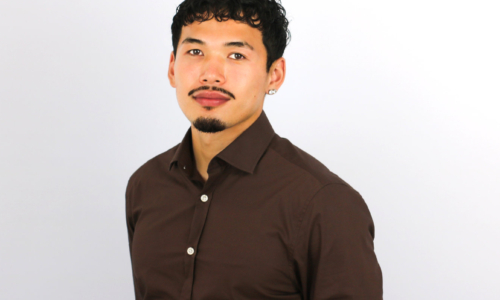 A young man with curly dark hair and facial hair smiles at the camera while wearing a dark brown button-up shirt with white buttons against a light gray background.