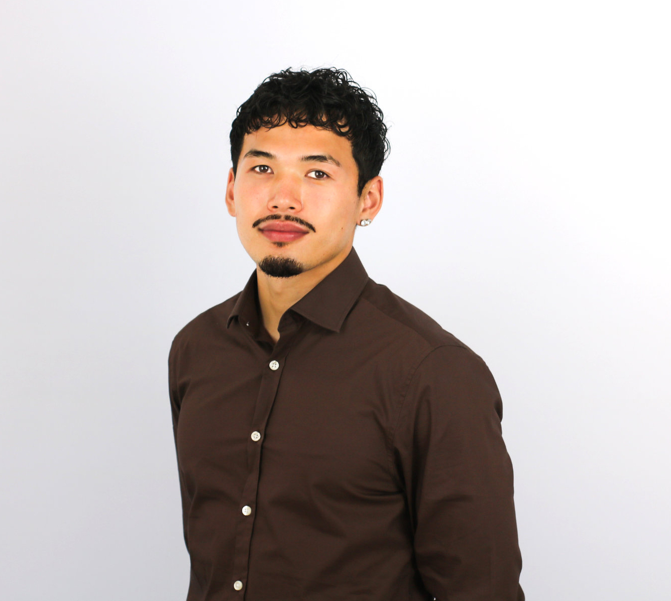 A young man with curly dark hair and facial hair smiles at the camera while wearing a dark brown button-up shirt with white buttons against a light gray background.