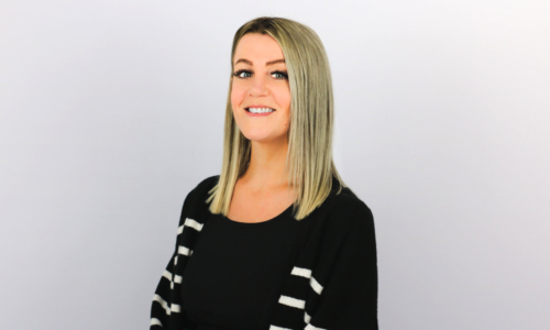 A smiling woman with shoulder-length blonde hair wearing a black sweater with white horizontal stripes on the sleeves, photographed against a light gray background in what appears to be a professional headshot style.