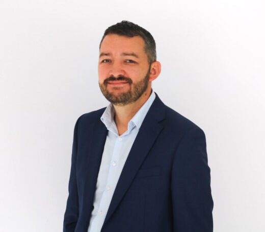A professional headshot of a smiling man with dark hair and a beard, wearing a navy blue blazer over a light blue dress shirt against a white background.