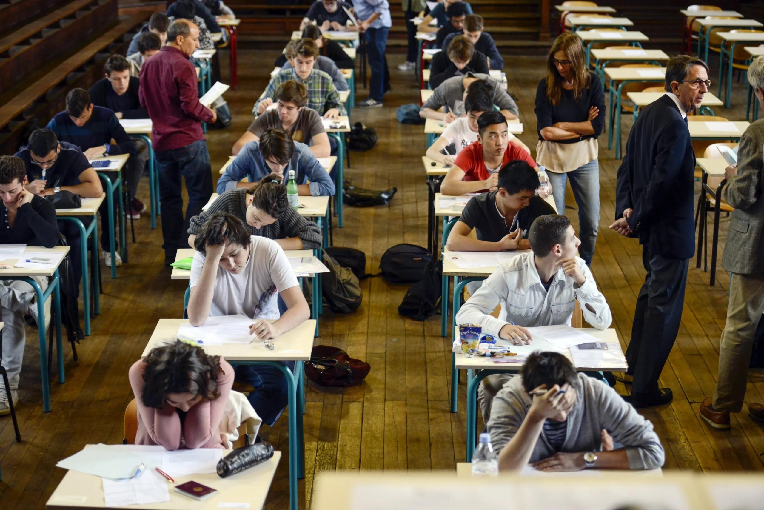 Students take an exam in a large hall with wooden floors, sitting at individual desks arranged in rows while proctors in business attire supervise by walking between the aisles.