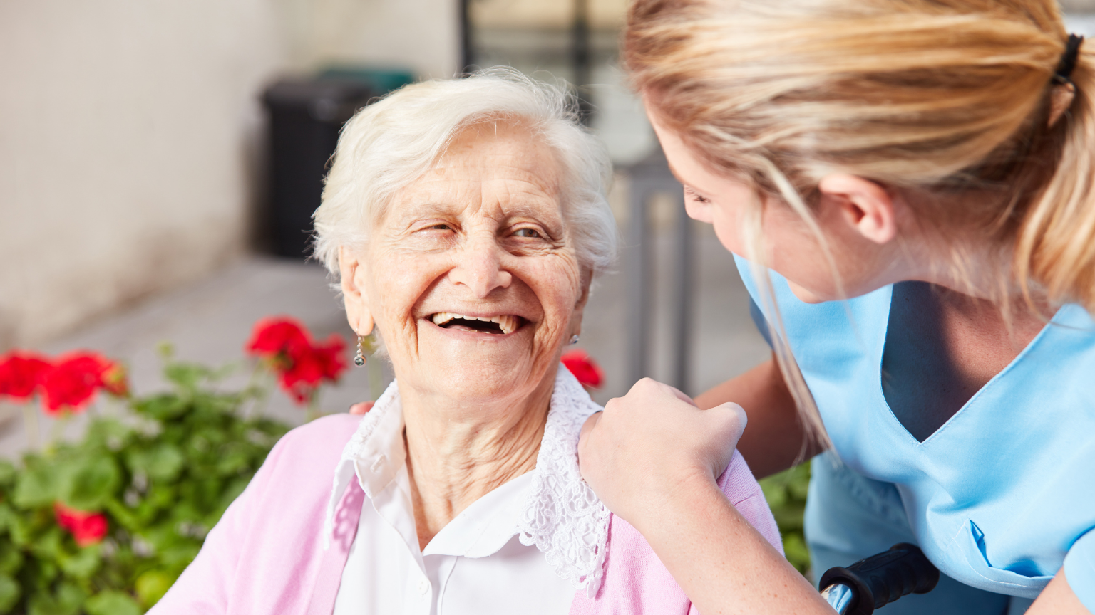An elderly woman with white hair and a bright smile sits next to a healthcare worker in blue scrubs who has her hand gently placed on the woman's shoulder, with red flowers visible in the background.