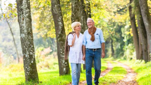 A mature couple walks arm-in-arm along a tree-lined path in a sunny park or wooded area, with the woman wearing a white top and jeans while the man wears a light blue shirt with a brown sweater draped over his shoulders.
