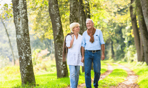 A mature couple walks arm-in-arm along a tree-lined path in a sunny park or wooded area, with the woman wearing a white top and jeans while the man wears a light blue shirt with a brown sweater draped over his shoulders.