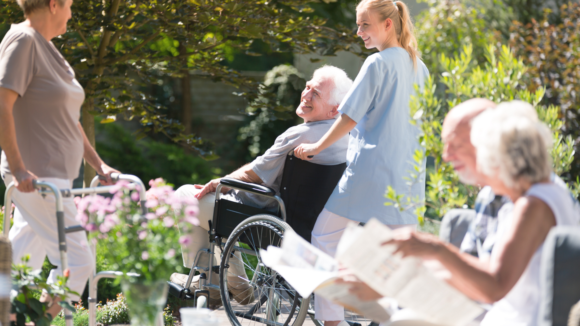 A healthcare worker in light blue scrubs assists an elderly man in a wheelchair while other seniors enjoy time outdoors in a sunny garden setting with blooming flowers.