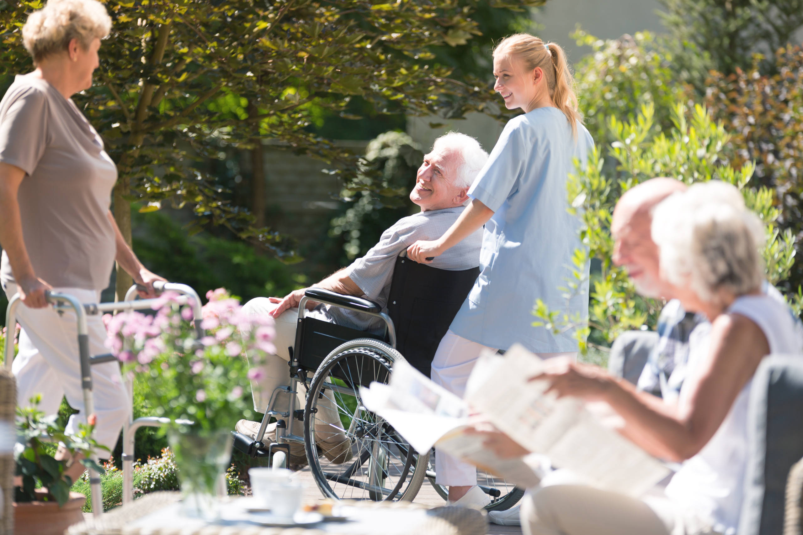 A healthcare worker in light blue scrubs assists an elderly man in a wheelchair while other seniors enjoy time outdoors in a sunny garden setting with blooming flowers.