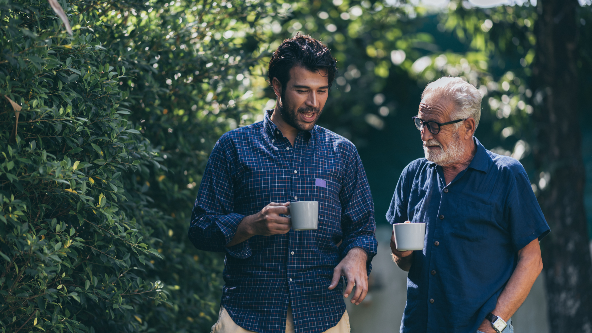 Two men of different generations walk together outdoors holding coffee cups, with the younger man in a plaid shirt and the older man with gray hair and glasses wearing a blue shirt, surrounded by lush green foliage.