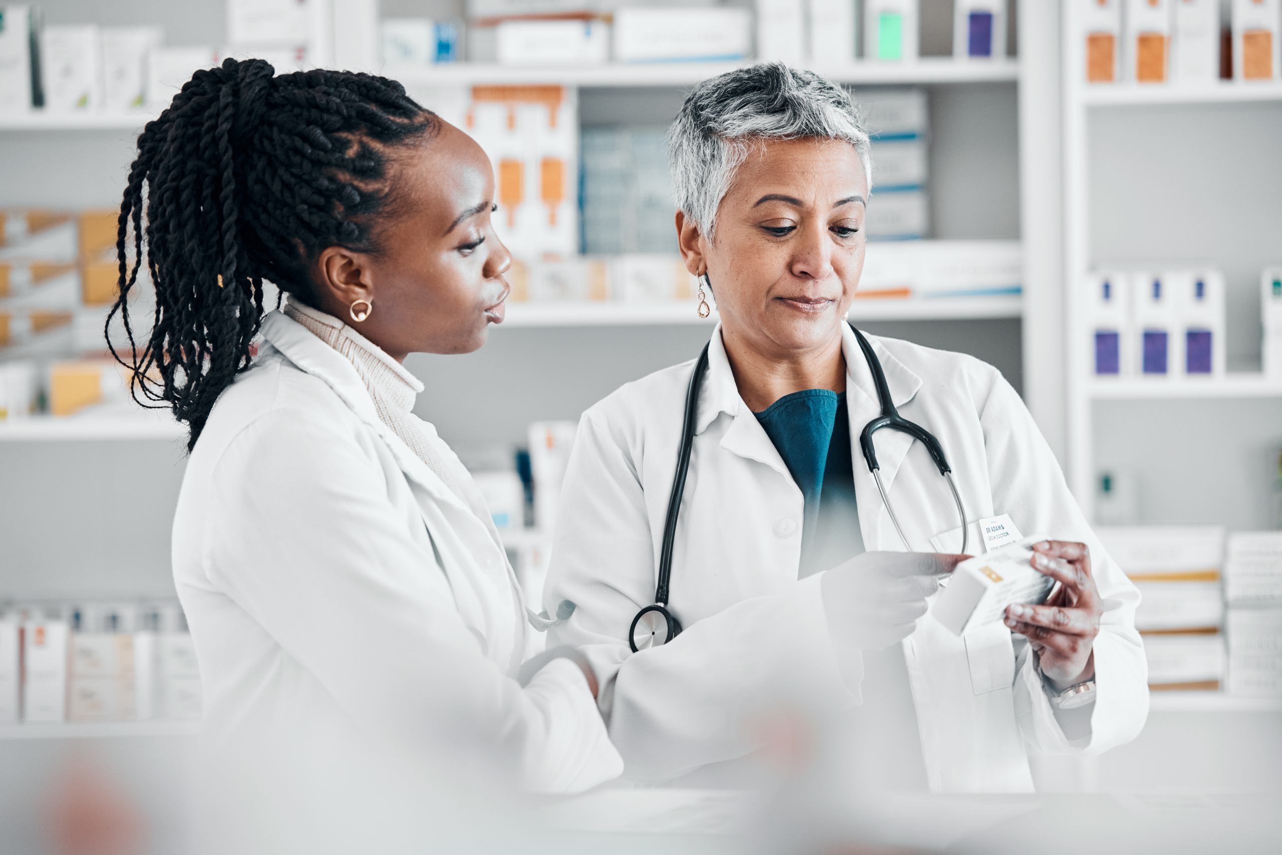 Two female healthcare professionals in white coats discuss medication in a pharmacy, with one wearing a stethoscope and holding a prescription bottle while shelves of medications are visible in the background.