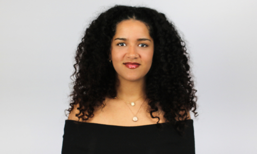 A woman with long, curly black hair smiles at the camera while wearing a black off-shoulder top and layered necklaces against a light gray background.