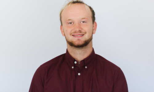 A smiling man with blonde hair and a beard wearing a burgundy button-down shirt against a white background in what appears to be a professional headshot photo.