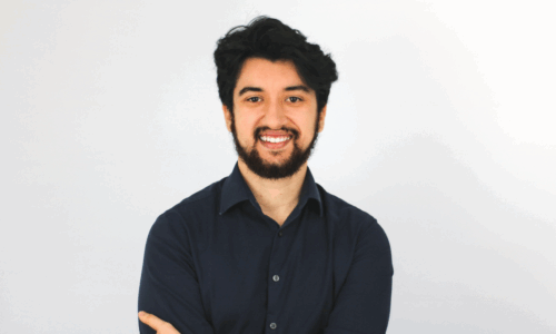 A smiling young man with dark curly hair and a beard stands with arms crossed, wearing a navy blue button-up shirt and a watch, against a white background.