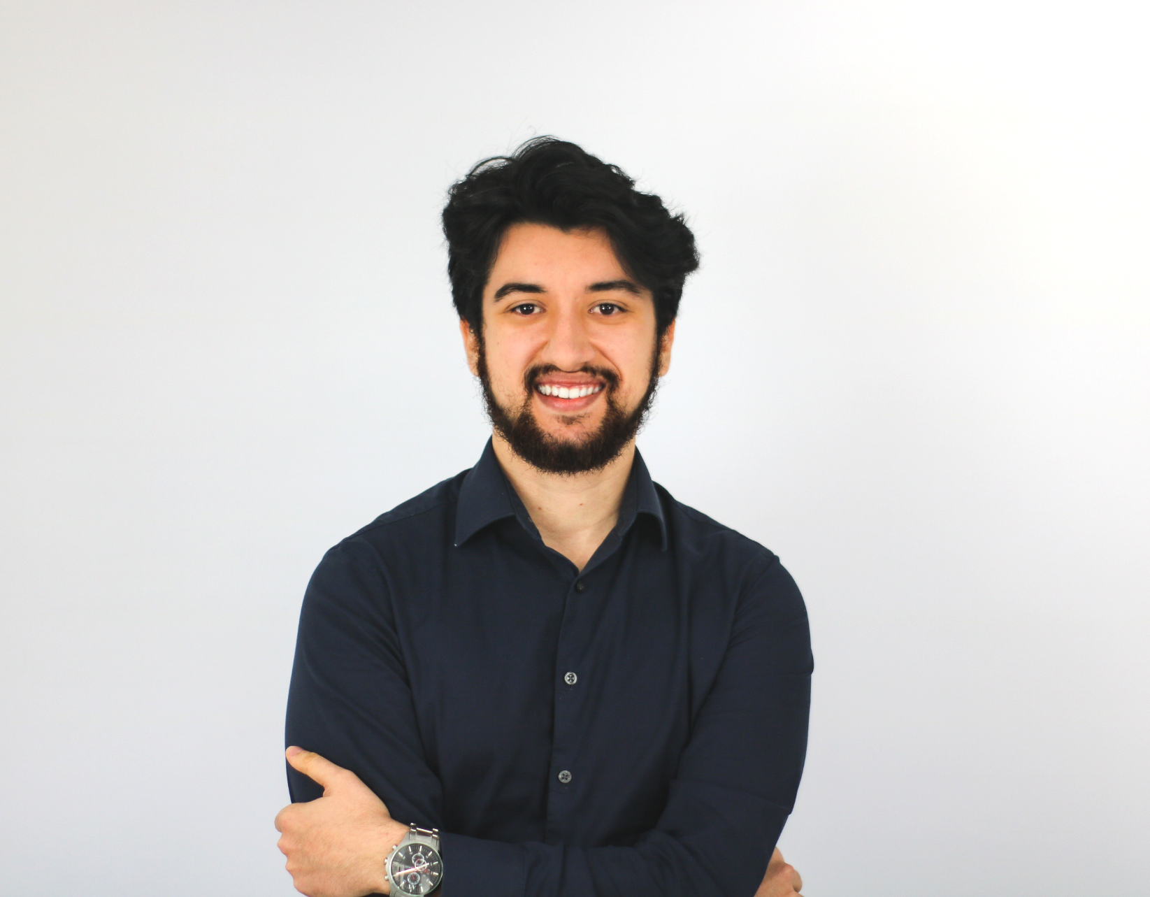 A smiling young man with dark curly hair and a beard stands with arms crossed, wearing a navy blue button-up shirt and a watch, against a white background.
