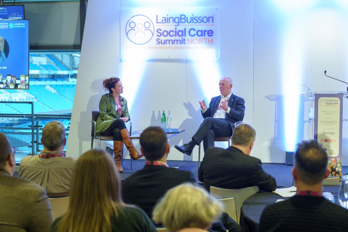 A panel discussion at the LaingBuisson Social Care Summit North featuring two speakers on stage - a woman in a green patterned jacket and a man in a dark suit - addressing an audience of professionally dressed attendees in what appears to be a conference venue.