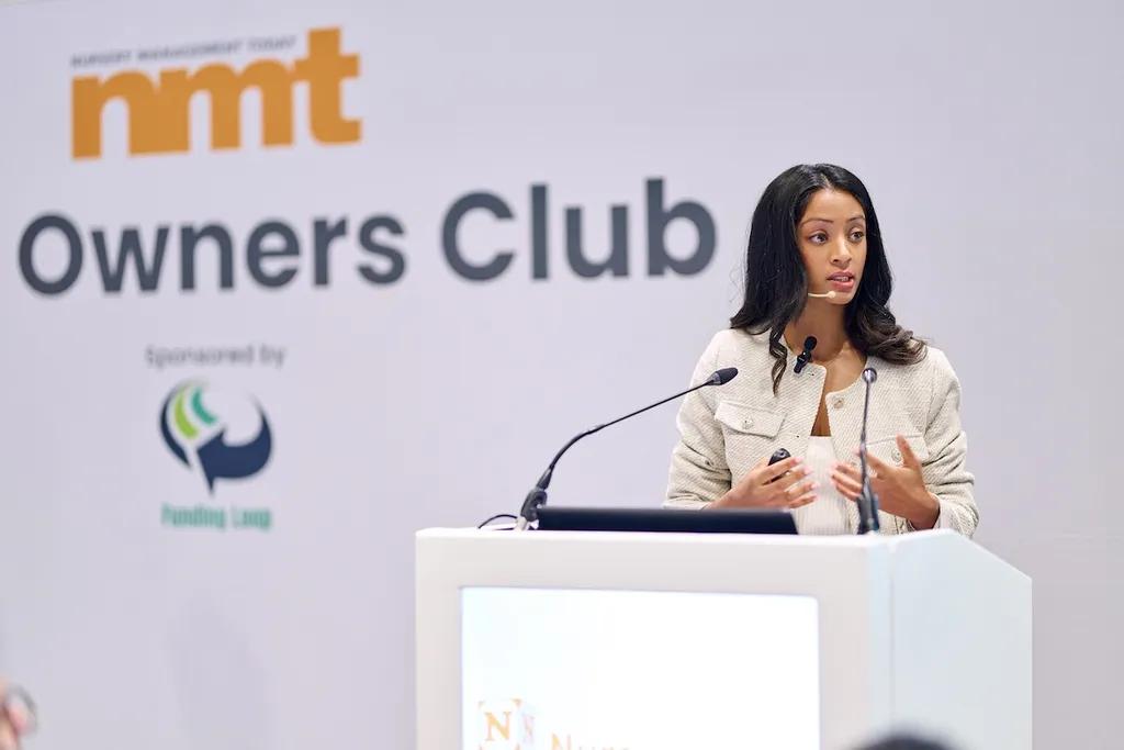 A woman in a beige blazer speaks at a podium during an RMT Owners Club event, with her hands gesturing as she addresses the audience.