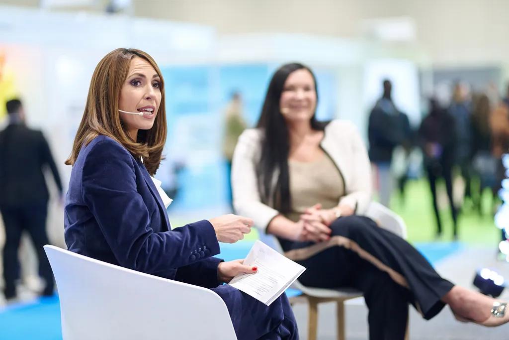 Two professional women are seated on white chairs in what appears to be a conference or event setting, with one woman in a navy blazer holding papers and speaking while wearing a microphone headset, and another woman in light-colored clothing listening in the background as attendees move around the venue.