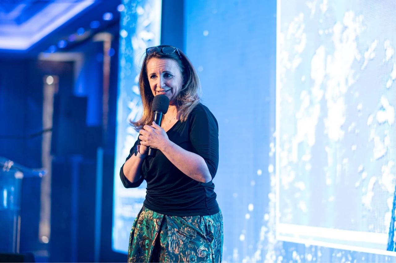 A woman in a black top and patterned skirt speaks into a microphone on stage, with blue lighting and a projection screen visible in the background.