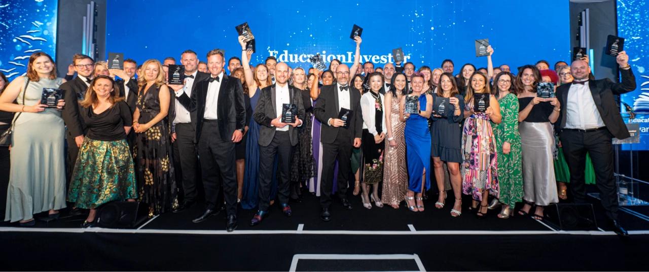 A large group of formally dressed award recipients poses together on stage at an educational investment awards ceremony, with many holding trophy plaques and wearing evening gowns and tuxedos against a blue backdrop.