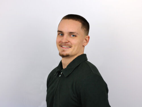 A man with short dark hair and a goatee smiles at the camera while wearing a dark green collared shirt against a light gray background.