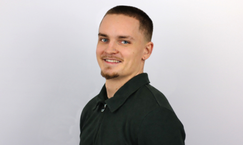 A man with short dark hair and a goatee smiles at the camera while wearing a dark green collared shirt against a light gray background.