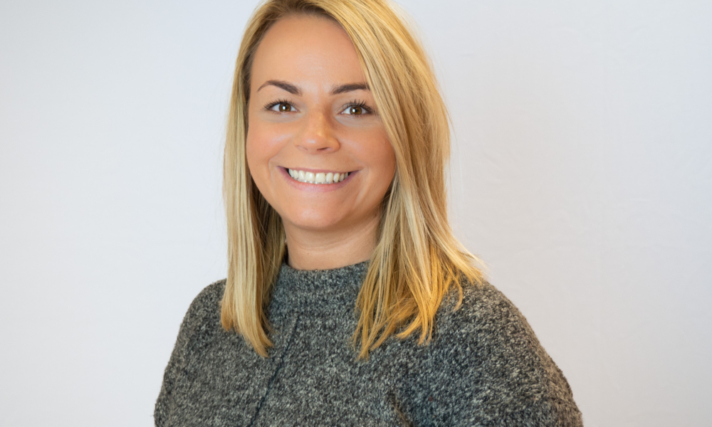 A smiling woman with blonde hair wearing a gray knitted sweater poses against a white background in what appears to be a professional headshot or portrait.