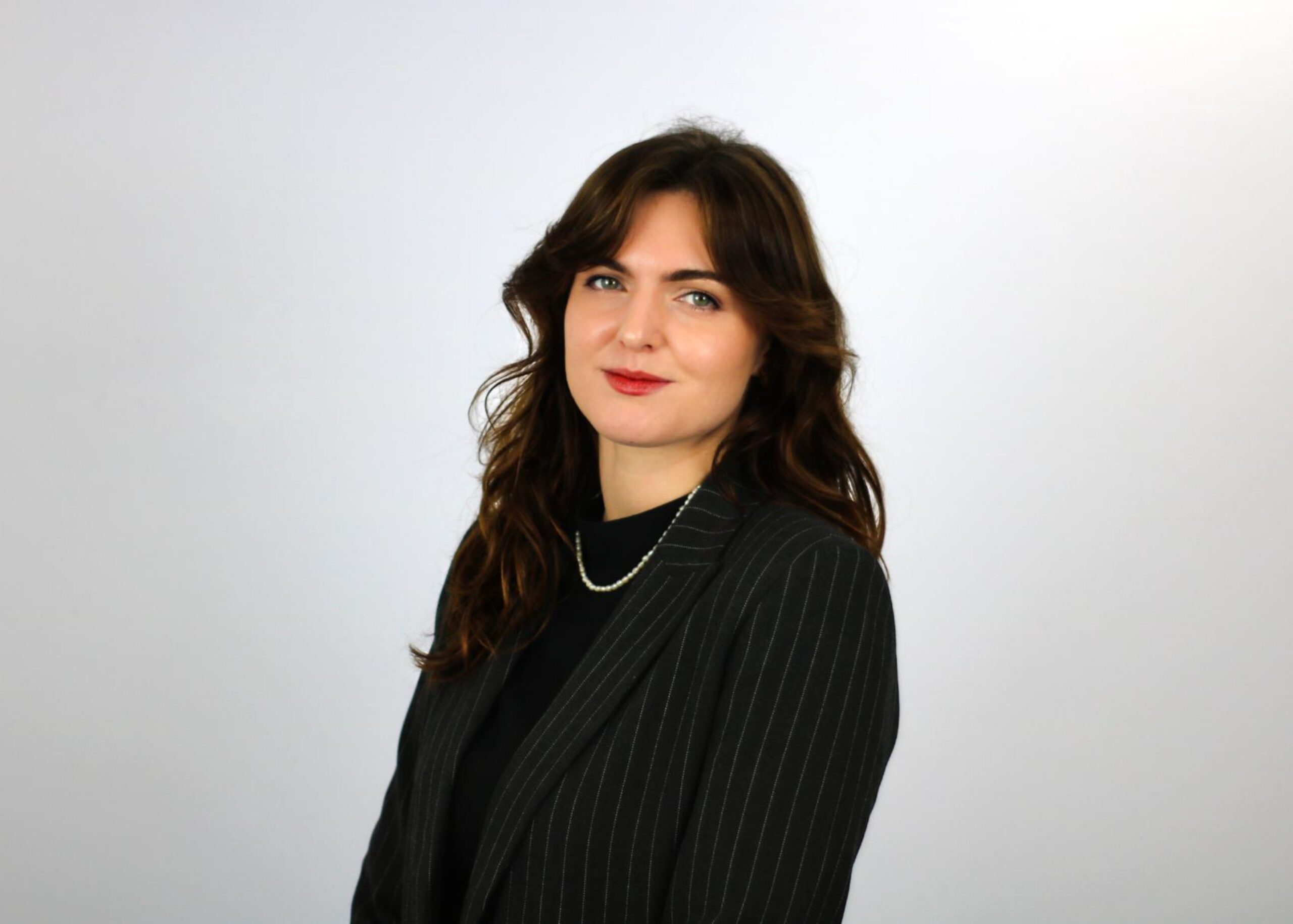 A woman with shoulder-length brown hair wearing a black pinstriped blazer and pearl necklace poses against a light gray background in what appears to be a professional headshot.