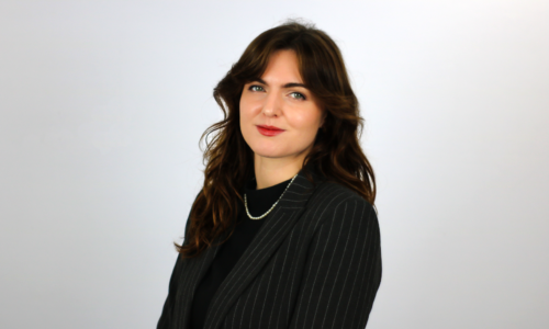 A woman with shoulder-length brown hair wearing a black pinstriped blazer and pearl necklace poses against a light gray background in what appears to be a professional headshot.