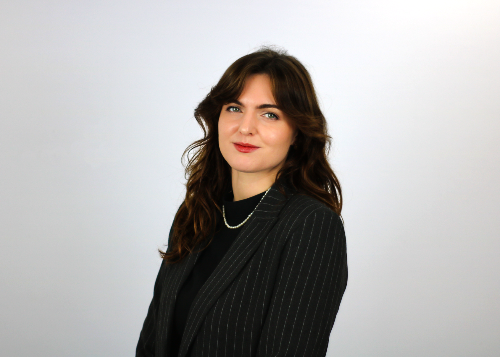 A woman with shoulder-length brown hair wearing a black pinstriped blazer and pearl necklace poses against a light gray background in what appears to be a professional headshot.