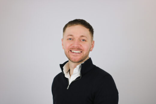 A smiling man with short brown hair and facial stubble wearing a black quarter-zip pullover over a white collared shirt, photographed against a plain light gray background.