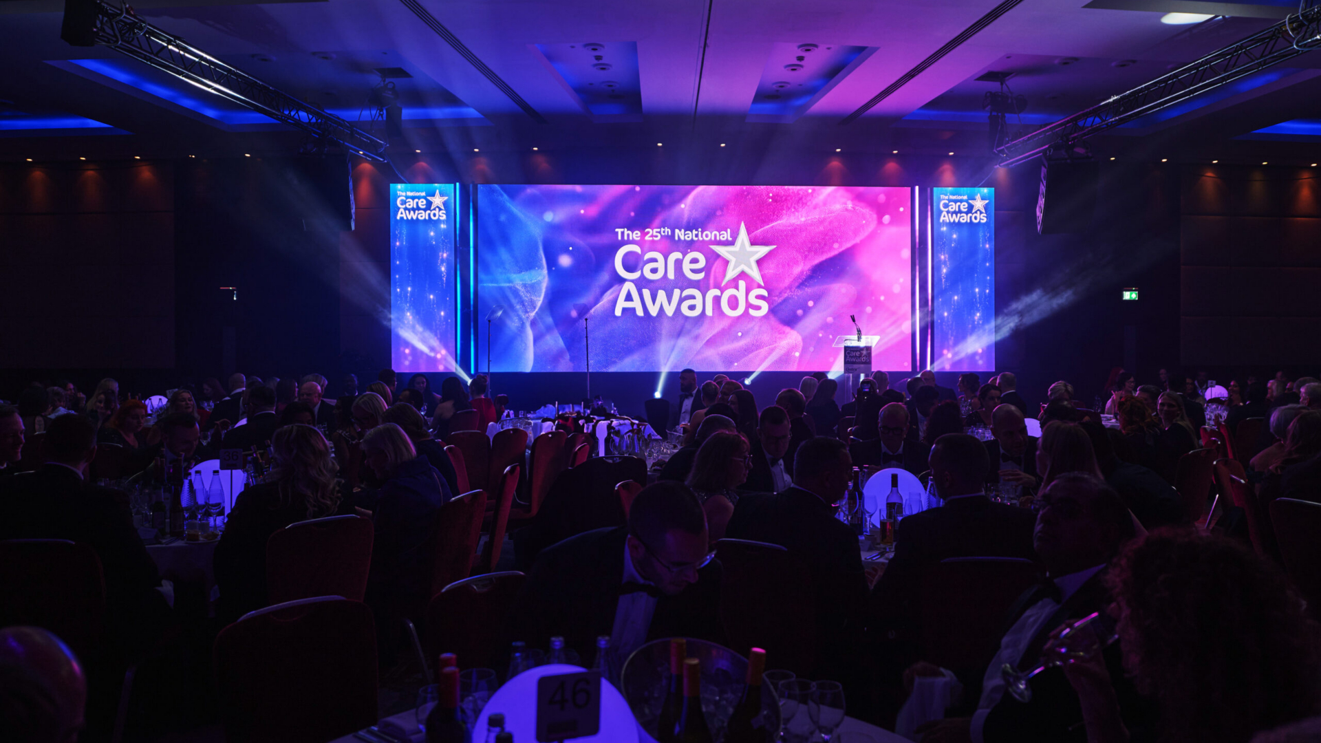 A large banquet hall filled with attendees at round tables during the 25th National Care Awards ceremony, featuring a brightly lit stage with purple and blue lighting displaying the Care Awards logo and branding on multiple screens.