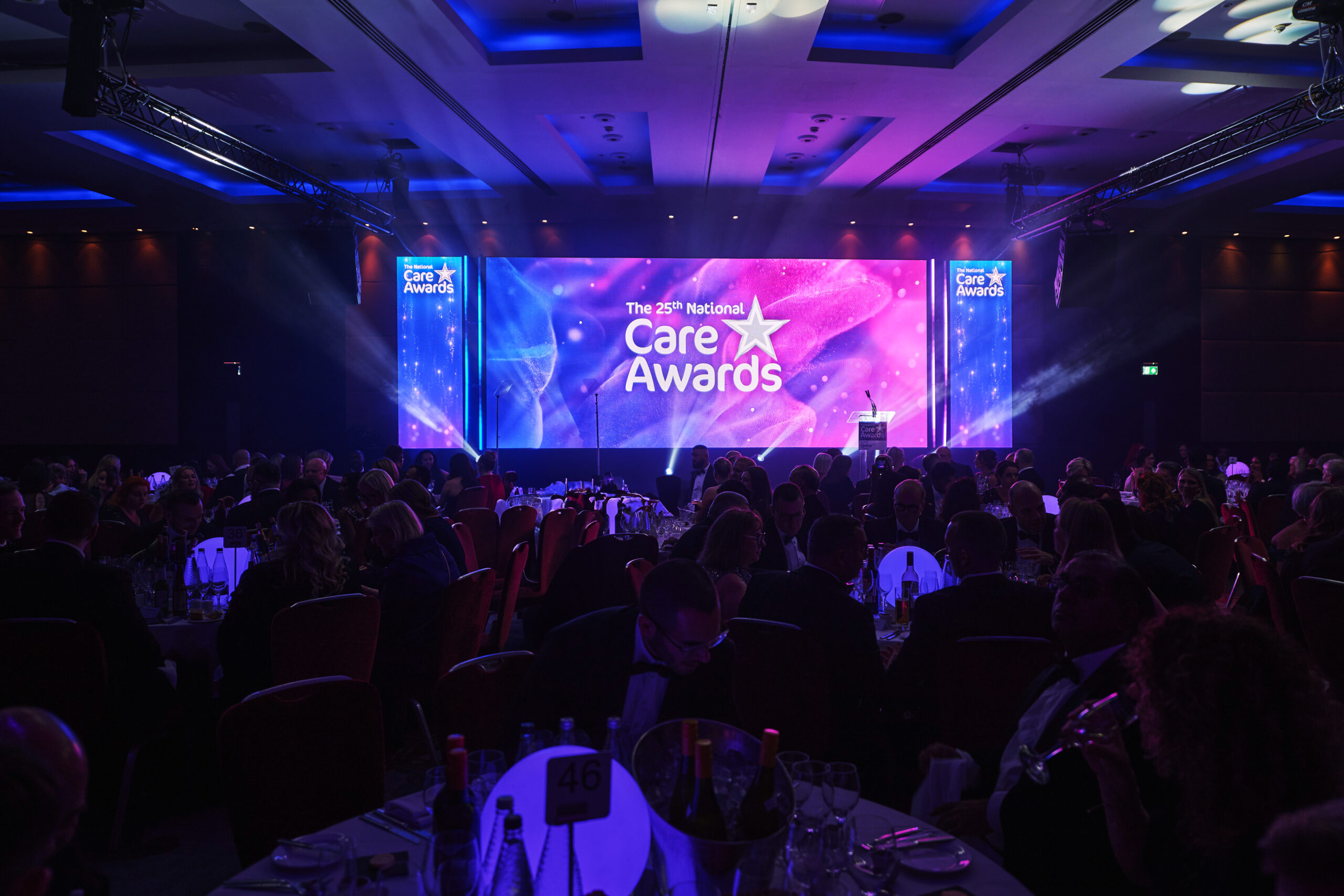 A large banquet hall filled with attendees at round tables during the 25th National Care Awards ceremony, featuring a brightly lit stage with purple and blue lighting displaying the Care Awards logo and branding on multiple screens.