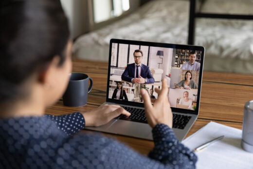 A person in a polka dot shirt participates in a video conference call on a laptop, with one hand raised while five colleagues appear in a grid layout on the screen.
