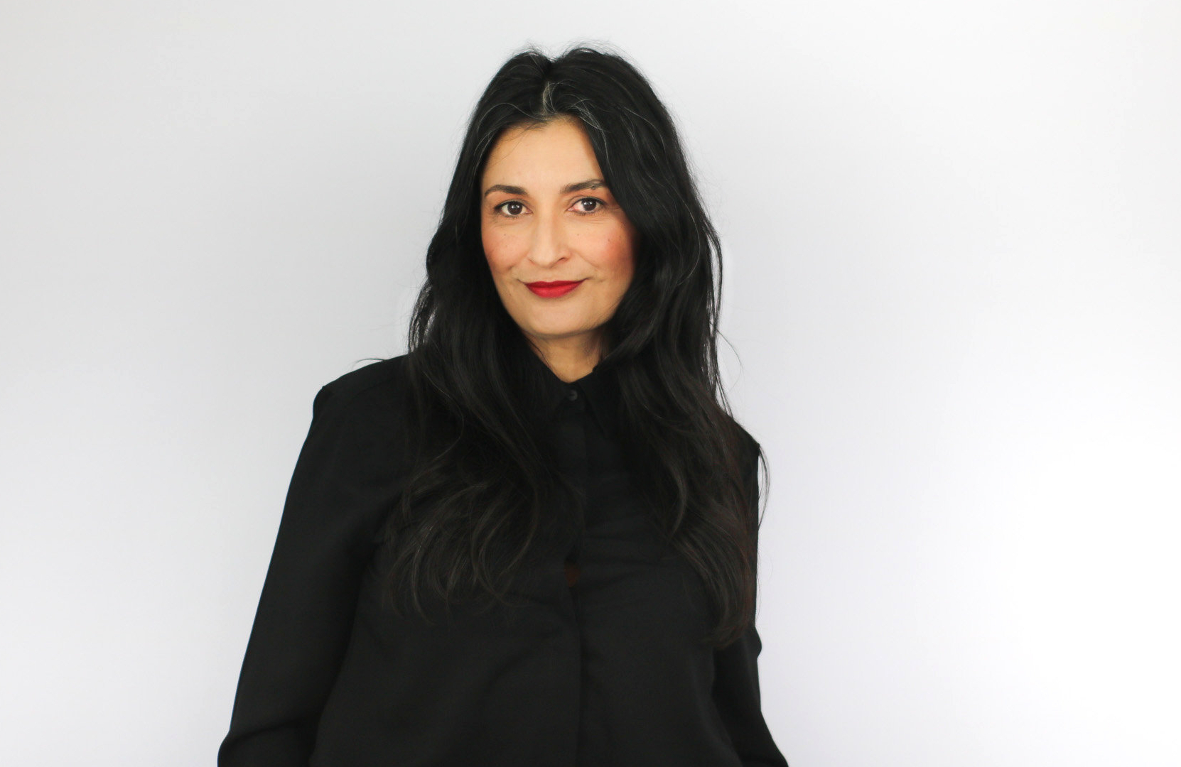 A woman with long dark hair wearing a black hoodie smiles at the camera against a white background, appearing to be in a professional headshot or portrait setting.