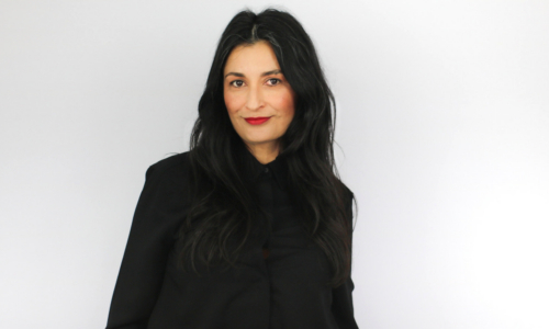 A woman with long dark hair wearing a black hoodie smiles at the camera against a white background, appearing to be in a professional headshot or portrait setting.