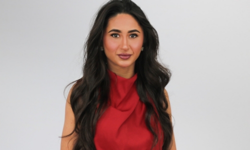A woman with long dark wavy hair wearing a red sleeveless top poses against a light gray background in what appears to be a professional portrait or headshot.