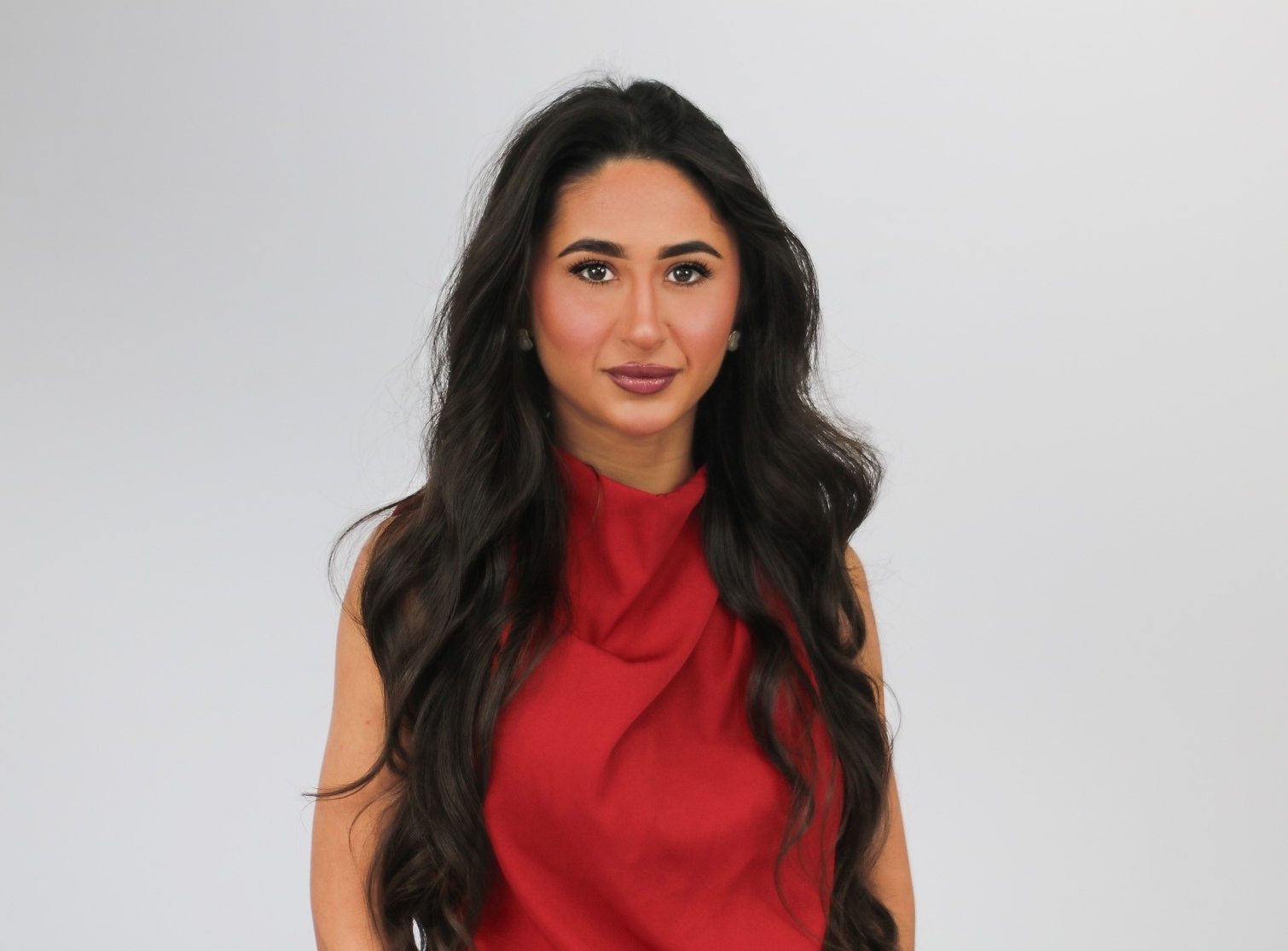 A woman with long dark wavy hair wearing a red sleeveless top poses against a light gray background in what appears to be a professional portrait or headshot.