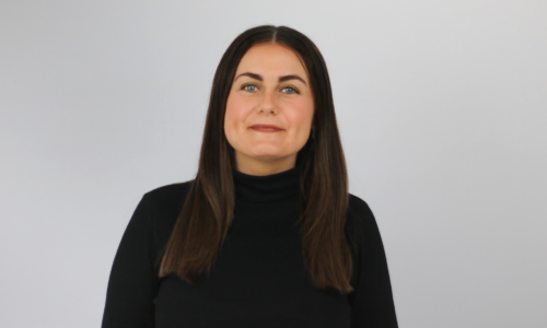 A woman with long brown hair and blue eyes wears a black turtleneck sweater while looking upward and to the side with a slight smile, photographed against a plain white background.