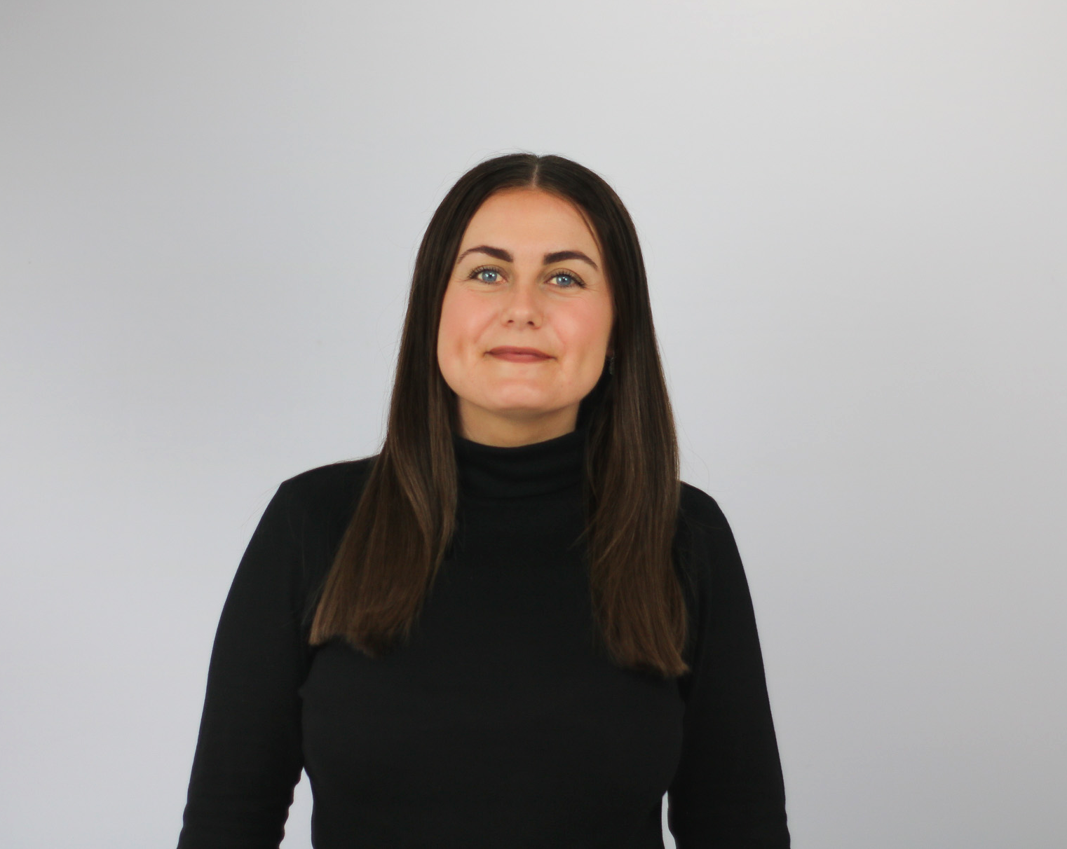 A woman with long brown hair and blue eyes wears a black turtleneck sweater while looking upward and to the side with a slight smile, photographed against a plain white background.