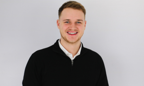 A young man with short brown hair and facial stubble smiles broadly at the camera, wearing a black zip-up sweater over a white collared shirt against a plain gray background.