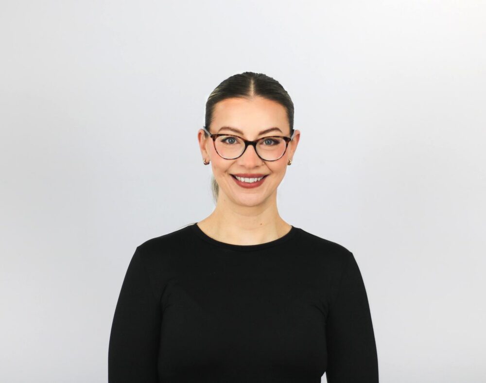 A woman with dark hair pulled back wearing black-rimmed glasses and a black long-sleeved top, smiling at the camera against a light gray background.