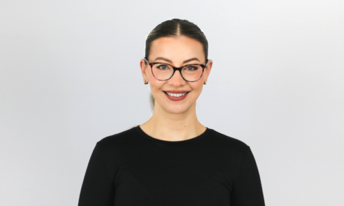 A woman with dark hair pulled back wearing black-rimmed glasses and a black long-sleeved top, smiling at the camera against a light gray background.