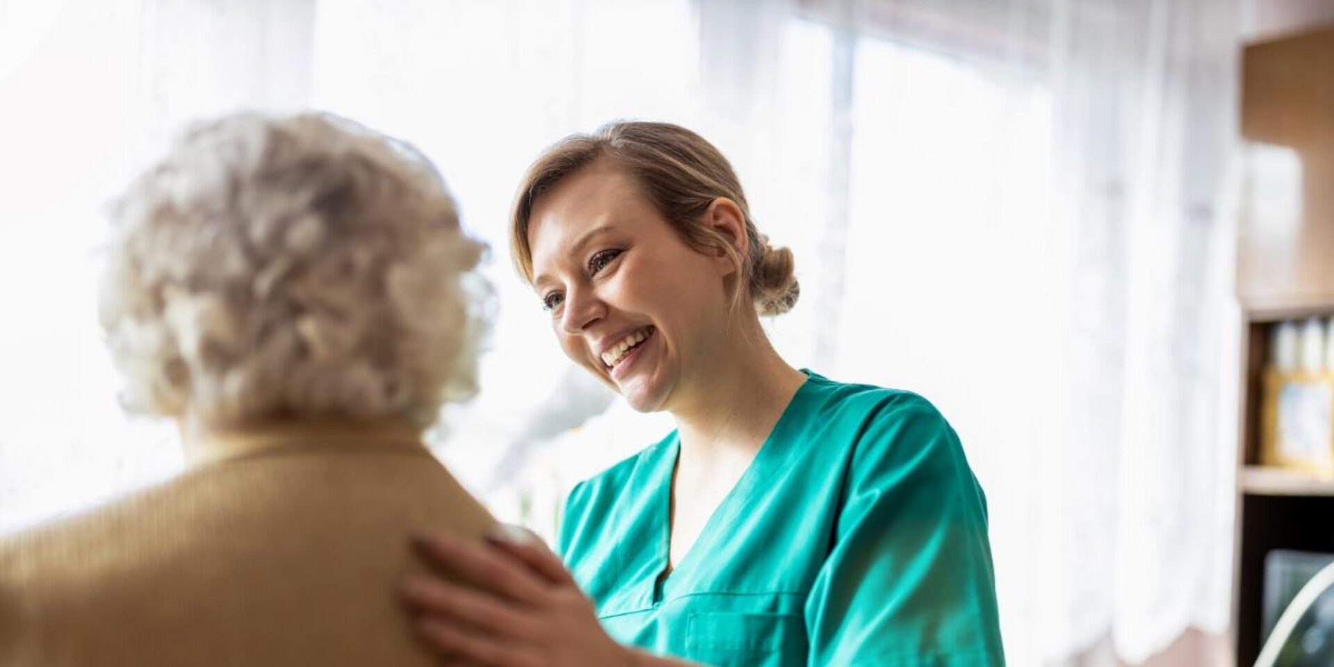 A smiling healthcare worker in green scrubs speaks with an elderly patient with gray curly hair in a bright, windowed room.