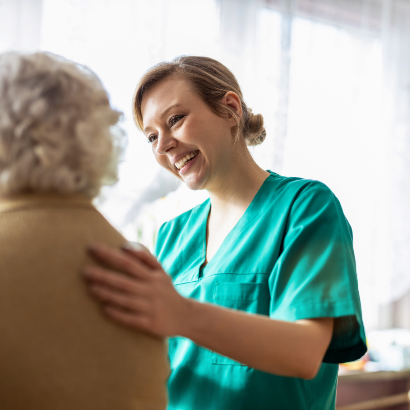A smiling healthcare worker in green scrubs provides compassionate care to an elderly patient with curly gray hair in a bright medical facility.